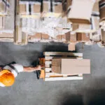 Um trabalhador em um armazém manobrando um carrinho com pallets descartáveis e caixas de papelão. O ambiente é organizado e mostra prateleiras lotadas de mercadorias.
